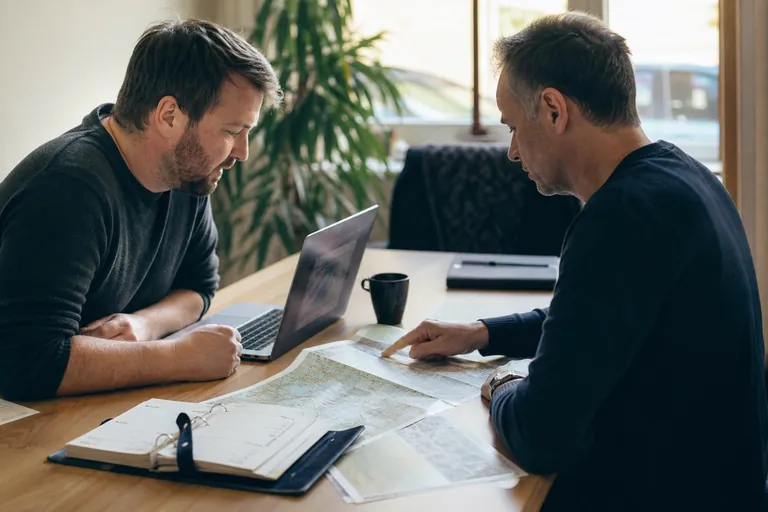 Zwei Fahrlehrer vergleichen Papierunterlagen mit einer Tabelle auf einem Laptop